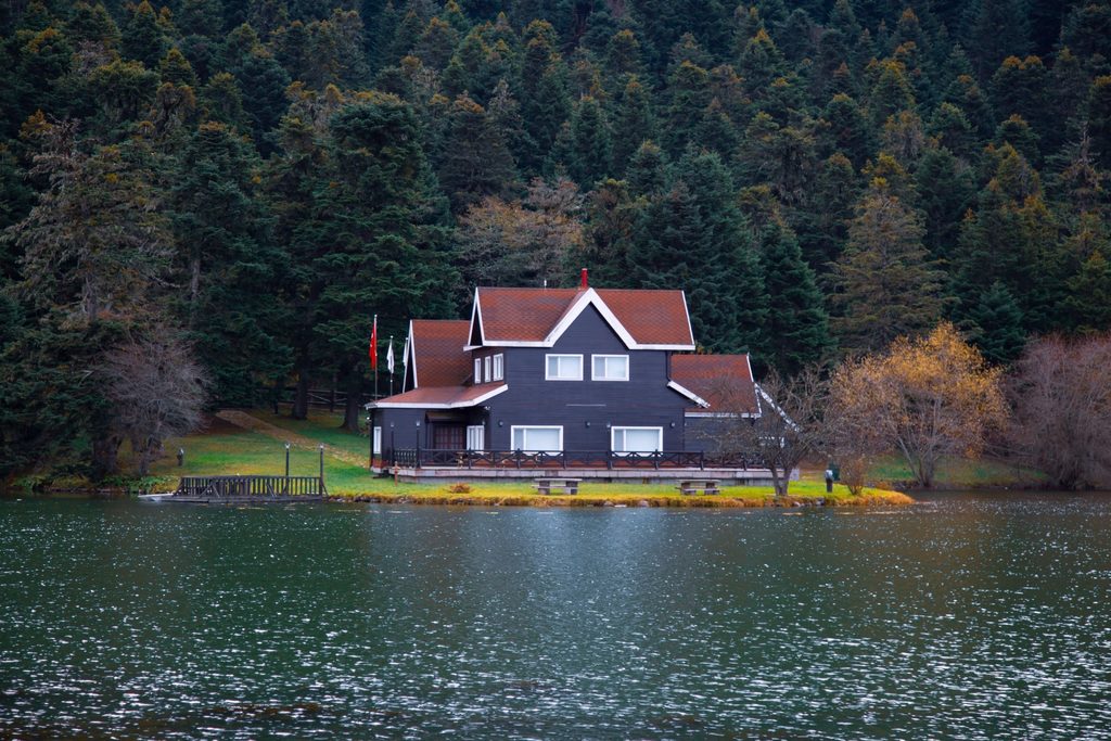 A wooden house with a red roof on the edge of a calm lake surrounded by a dense evergreen forest